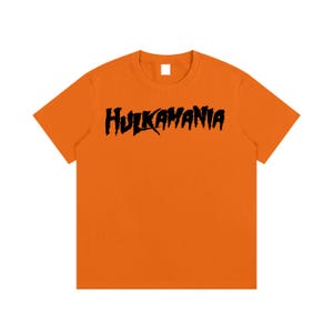May include: A bright orange short-sleeved t-shirt featuring the word HULKAMANIA printed in a bold, black, jagged, distressed-style font across the center of the chest. The garment has a standard crew neckline and a solid, vibrant orange color.