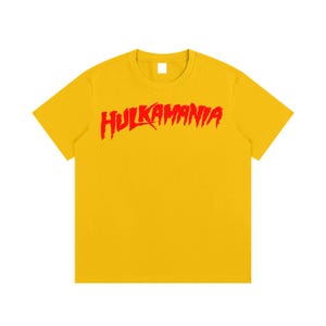May include: A bright yellow short-sleeved t-shirt featuring the word HULKAMANIA printed in a bold, jagged red font across the chest. The garment has a classic crew neckline and a solid color design, providing a simple and clean aesthetic.