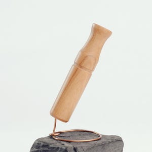 Puede incluir: Un objeto de madera clara, posiblemente una herramienta o un artículo decorativo, se exhibe sobre una piedra gris oscura. Un anillo de alambre de cobre sostiene la pieza de madera. El fondo es blanco liso.