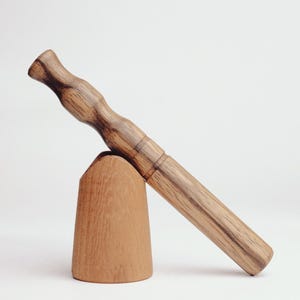 Puede incluir: Un juego de mortero y mazo de madera. El mazo tiene forma larga y cilíndrica con un mango redondeado. El mortero es un cuenco de madera pequeño en forma de cono. El conjunto es de madera marrón claro.