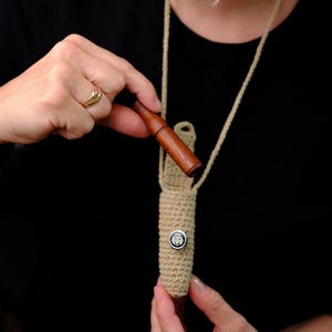 Op de afbeelding: Een houten pijp wordt in een beige gehaakte hoes met een knoopsluiting geplaatst. De hoes is bevestigd aan een beige koordketting. De pijp is donkerbruin van kleur en de hoes is lichtbeige.