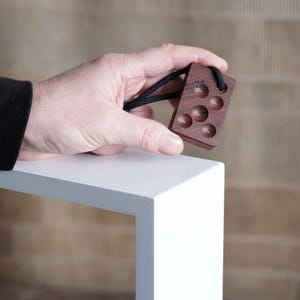 Op de afbeelding: Een klein, rechthoekig houten object met zes cirkelvormige inkepingen en een zwarte koordlus. Het hout heeft een rijke, donkerbruine kleur. Het object wordt tegen een neutrale achtergrond gehouden.