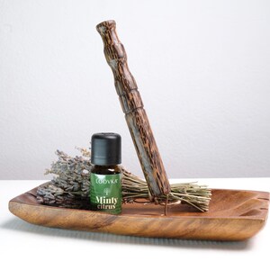 Peut inclure: Un plateau rectangulaire en bois contenant un bouquet de lavande séchée, un petit flacon en verre brun avec un bouchon noir étiqueté "LOOVKA Minty Citrus" et un long bâton en bois brun texturé dressé dans un support métallique.