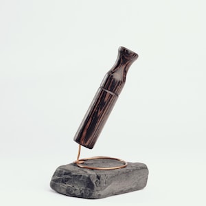 Op de afbeelding: Een donkerbruin houten object met een cilindrisch lichaam en een uitlopende bovenkant, tentoongesteld op een koperdraadstandaard bovenop een grijze stenen basis. De houtnerf is zichtbaar. Het object is lichtjes schuin.