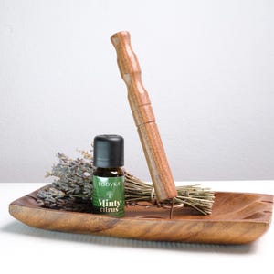 May include: A rectangular wooden tray holding a small brown glass bottle with a black cap labeled Loovka Minty Citrus, a bundle of dried lavender stems, and a tall, cylindrical wooden pestle-like tool standing upright in the center.
