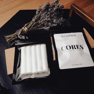 Może przedstawiać: Kompozycja z suszoną lawendą, opakowaniem białych cylindrycznych rdzeni i paczką z napisem "CORES for loovka air diffuser". Przedmioty znajdują się na czarnej powierzchni z drewnianym tłem.