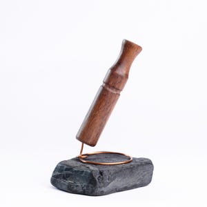 Könnte beinhalten: Ein handgefertigtes Holzobjekt mit dunkelbraunem Finish, das auf einem dunkelgrauen Steinsockel ruht. Ein Kupferdrahtring und eine Haltestruktur halten das Holzstück. Der Hintergrund ist weiß.