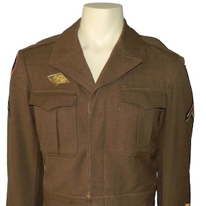 Może przedstawiać: Brązowa kurtka wojskowa z wykładanym kołnierzem, dwiema kieszeniami na piersi i naszywkami na ramionach. Kurtka ma złote i czarne paski na rękawach oraz złoty emblemat na piersi. Kurtka jest prezentowana na manekinie.