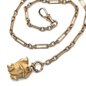 May include: A gold-toned metal chain featuring elongated rectangular links connected by smaller circular links. The chain includes a swivel clasp and a spring ring, with a carved, off-white, textured animal-shaped pendant attached to the end.