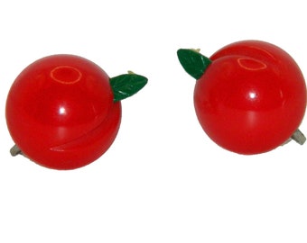 May include: A pair of bright red, spherical fruit-shaped earrings, each featuring a small, glossy green leaf attached to the side. The earrings have a smooth, polished finish and a rounded design that mimics the appearance of a small red apple.