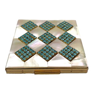 May include: A square compact mirror case featuring a gold-tone metal frame and a top surface inlaid with a checkerboard pattern of iridescent white mother-of-pearl tiles and eight diamond-shaped clusters of small, bright light blue rhinestones.