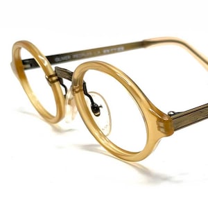 May include: Oliver Peoples eyeglasses featuring round, translucent amber-toned acetate frames and metallic temples with an intricate engraved scrollwork pattern. The design includes adjustable nose pads and curved temple tips for a secure fit.