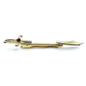 May include: A gold-toned tie clip in the shape of a sword. The sword features a red gemstone at the hilt and a decorative handle. The clip is designed to secure a tie and adds a touch of elegance.