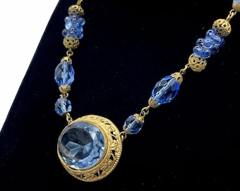 May include: A vintage necklace featuring a large, light blue faceted pendant set in a gold-toned filigree frame. The necklace has matching blue glass beads and ornate gold-toned metal accents. The necklace is displayed against a black background.