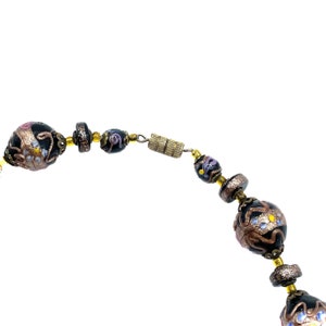 May include: A beaded necklace featuring black, gold, and bronze beads. The beads are decorated with a floral pattern and blue and yellow accents. The necklace is secured with a gold-coloured clasp.