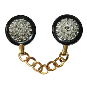 May include: A pair of black and silver round buttons connected by a gold chain. The buttons have a decorative design with a silver center.