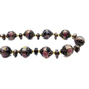 May include: A necklace with dark beads, each decorated with pink, blue, and yellow floral patterns. The beads are separated by smaller, dark brown beads and linked by a gold-toned chain. The background is plain white.