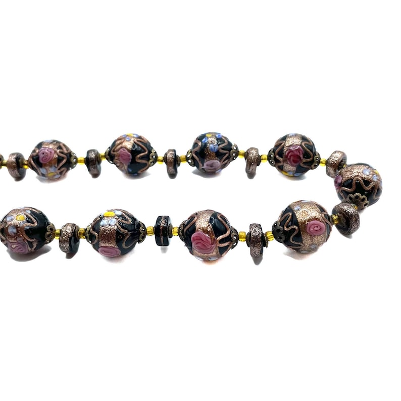 May include: A necklace with dark beads, each decorated with pink, blue, and yellow floral patterns. The beads are separated by smaller, dark brown beads and linked by a gold-toned chain. The background is plain white.