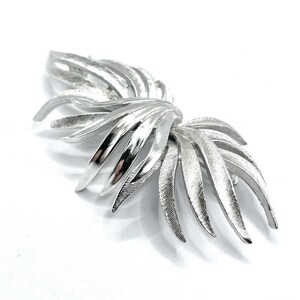 May include: A silver-toned brooch with a stylized leaf design. The brooch features multiple curved, textured leaves radiating outwards from a central point. The leaves have a brushed metal finish, creating a play of light and shadow.