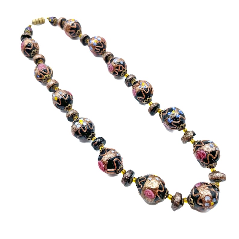 May include: A vintage necklace with black, gold, and pink glass beads. The beads have detailed floral patterns and are strung with gold-toned metal. The necklace is shown against a white backdrop.