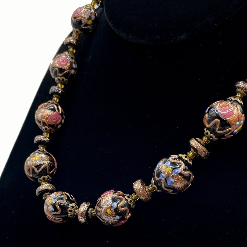 May include: A vintage necklace with large, round beads. The beads have intricate floral designs in pink, yellow, and blue on a black background. The beads are separated by smaller, gold-toned spacers, creating an elegant look.