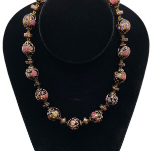 May include: A vintage necklace with large, round beads. The beads have a black base with pink, yellow, and blue floral patterns. Bronze-coloured spacers separate the beads. A unique and elegant necklace.