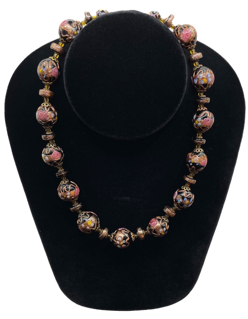 May include: A vintage necklace with large, round beads. The beads have a black base with pink, yellow, and blue floral patterns. Bronze-coloured spacers separate the beads. A unique and elegant necklace.