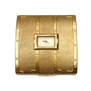 Op de afbeelding: Een goudkleurig metalen compact met een scharnierend deksel en een klein, rond klokje met zwarte Romeinse cijfers. De compact heeft een getextureerd oppervlak en een decoratief ontwerp.