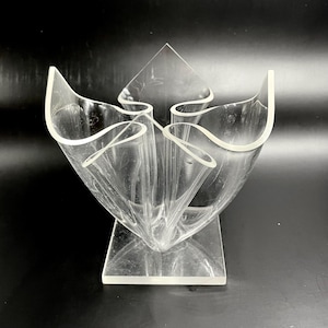 May include: Clear acrylic vase with a unique, abstract design. The vase features a square base and a series of curved, petal-like shapes that rise upwards. The transparent material allows light to pass through, creating interesting shadows.