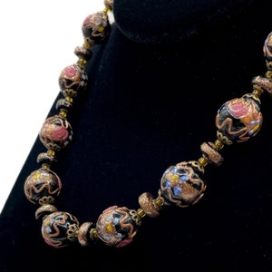 May include: A vintage necklace with large, round black beads, adorned with floral patterns in pink, gold, and blue. The beads are linked with smaller, textured gold-toned beads. The necklace is displayed against a black backdrop, showcasing its intricate design.