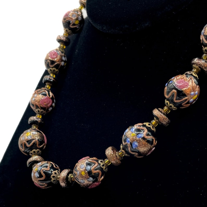 May include: A vintage necklace with large, round black beads, adorned with floral patterns in pink, gold, and blue. The beads are linked with smaller, textured gold-toned beads. The necklace is displayed against a black backdrop, showcasing its intricate design.