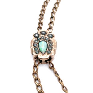 Peut inclure: Un collier doré avec un pendentif central à bord festonné, orné d'une opale iridescente en forme de poire au centre, entourée de petites pierres circulaires sombres. Le pendentif est fixé à une fine chaîne de couleur dorée.