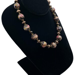 May include: A vintage necklace with black, round beads featuring floral designs in pink, yellow, and white. The necklace is displayed on a black velvet jewellery stand, showcasing its intricate details and craftsmanship.