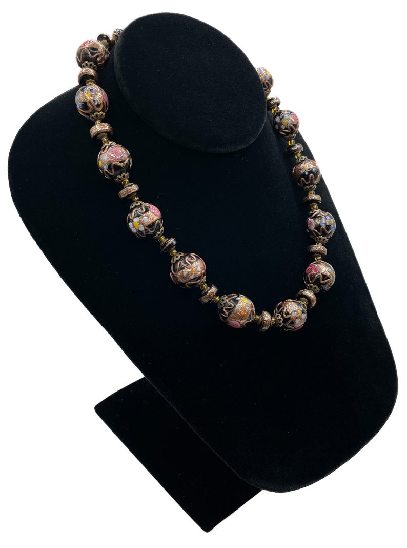 May include: A vintage necklace with black, round beads featuring floral designs in pink, yellow, and white. The necklace is displayed on a black velvet jewellery stand, showcasing its intricate details and craftsmanship.