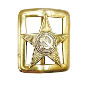 May include: A rectangular gold-coloured metal belt buckle featuring a prominent five-pointed star at its centre. The star is decorated with fine radiating lines and contains a circular emblem displaying a crossed hammer and sickle symbol.
