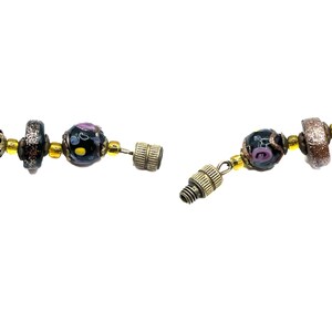 May include: A close-up of a necklace with a magnetic clasp. The necklace features black glass beads with colourful floral designs, gold-coloured beads, and a brown and gold bead. The clasp is a screw-type closure.