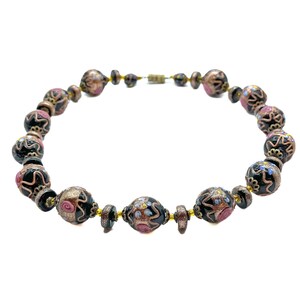 May include: A necklace with decorative beads in black, gold, and pink. The beads have intricate floral designs and are strung together with a gold-coloured chain and clasp. The necklace is displayed against a white background.