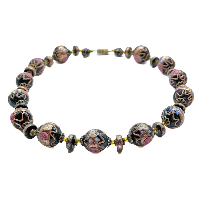May include: A necklace with decorative beads in black, gold, and pink. The beads have intricate floral designs and are strung together with a gold-coloured chain and clasp. The necklace is displayed against a white background.