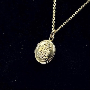 Puede incluir: Un collar con colgante de medallón ovalado dorado. El medallón presenta un diseño floral y cuelga de una delicada cadena. El fondo es de color oscuro y liso.