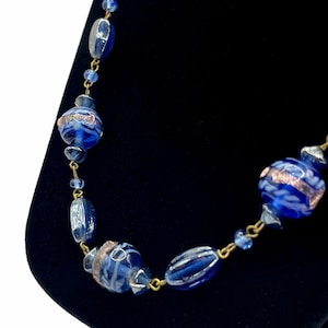 May include: A blue beaded necklace with gold-colored chain links. The necklace features various shapes of blue glass beads, including round and oval beads. Some beads have white and gold accents.