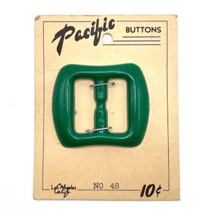 Op de afbeelding: Een glanzende, smaragdgroene rechthoekige riemgesp met afgeronde hoeken en een centrale balk, gemonteerd op een beige kartonnen displaykaart met het opschrift Pacific BUTTONS, Los Angeles Calif. NO 48 en 10 cents. De gesp heeft twee metalen pinnen.