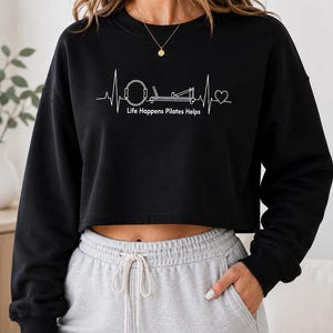 May include: A black long-sleeve cropped sweatshirt featuring a white graphic print of a heartbeat line that incorporates a Pilates ring and reformer machine, with the text "Life Happens Pilates Helps" and a heart symbol at the end of the line.