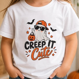 May include: A white short-sleeved t-shirt featuring a graphic of a ghost wearing an orange baseball cap and black sunglasses, holding a jack-o'-lantern. The text "CREEP IT Cute" is printed below in black and orange, surrounded by bats and stars.