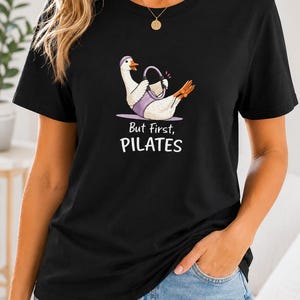 May include: A black short-sleeved t-shirt featuring a graphic of a white goose wearing a purple headband and using a purple pilates ring. Below the goose, the text reads "But First, PILATES" in white and black lettering. The shirt has a crew neckline.