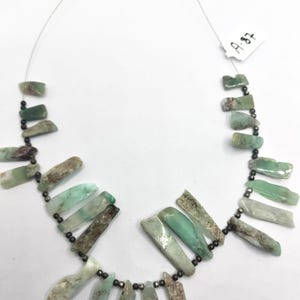 Può includere: Una collana composta da segmenti di pietra verde pallido, allungati e irregolari, intervallati da piccole perline metalliche scure. Le pietre presentano texture variegate nei toni del verde menta e salvia. È attaccata una piccola etichetta bianca A-87.
