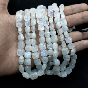 Puede incluir: Un hilo de cuentas de piedra lunar blancas, translúcidas y pulidas, con forma cuadrada y redondeada. Cada cuenta muestra una sutil adularescencia azul iridiscente que capta la luz, creando un brillo suave sobre la superficie de la piedra.