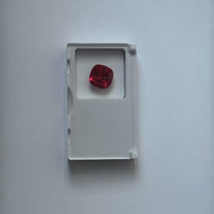 May include: A vibrant, cushion-cut, deep red gemstone is presented in a clear plastic case. The gem's square shape with rounded corners reflects light, highlighting its facets. The case is rectangular with a hinged lid.