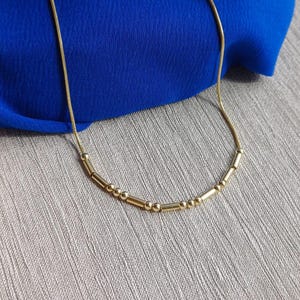 Pode incluir: Um delicado colar dourado com uma série de pequenas contas redondas e barras cilíndricas. O colar é exibido em um fundo de tecido azul, com um raminho de flores secas. A corrente é fina e flexível.