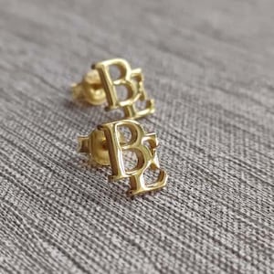May include: A pair of gold-colored stud earrings. Each earring features the letters "B" and "R" in a stylized, open design. The earrings are displayed on a textured, gray surface.