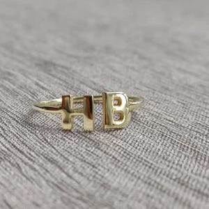 May include: A gold ring displaying the letters "H" and "B". The ring is set against a neutral, textured backdrop. The letters are in a sans-serif font.